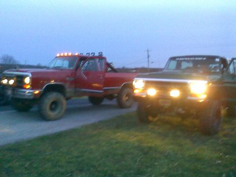 1979 Ford Bronco - Two modified pickup trucks are parked side-by-side in the twilight, their auxiliary lights illuminating the scene.