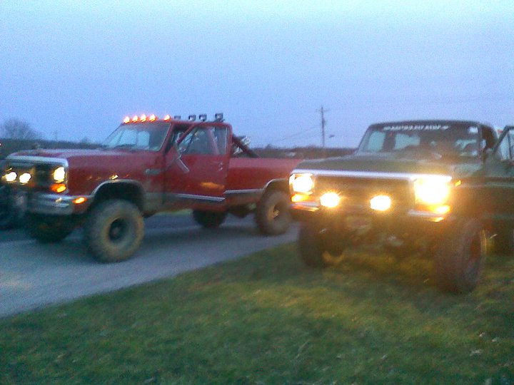 Two lifted trucks, one red and one dark colored, parked outside at dusk with their headlights on