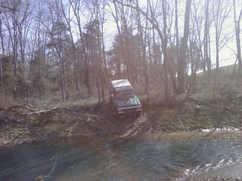 1979 Ford Bronco - A green 1979 Ford Bronco navigates a muddy bank next to a stream, surrounded by trees.
