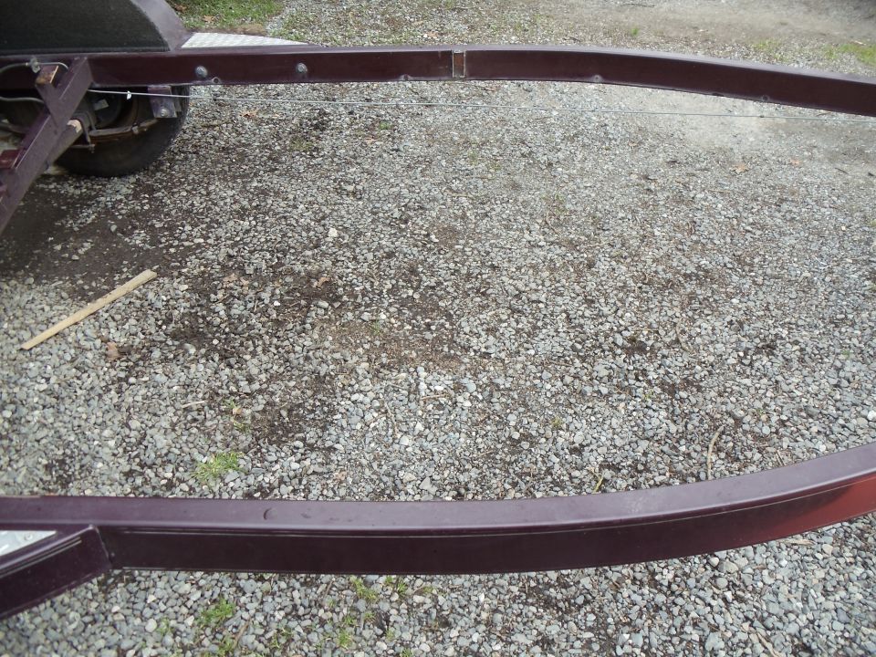 Dark maroon trailer frame rails over gravel with axle at left