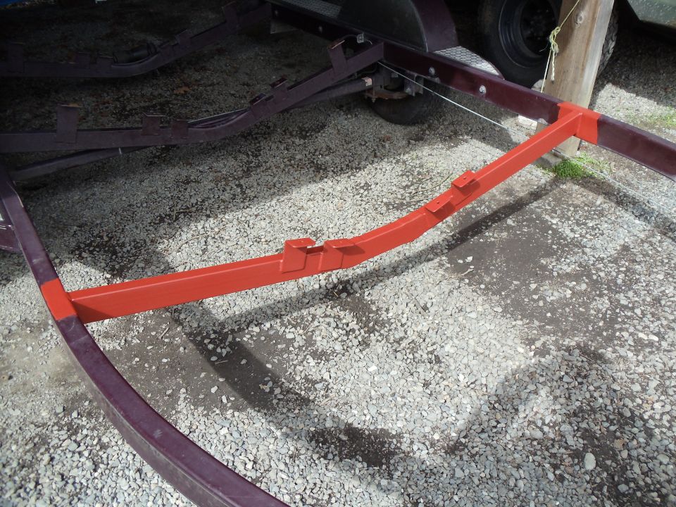 Orange-painted trailer crossmember on gravel beside a maroon boat trailer frame