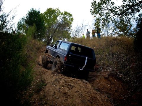 1995 Ford Bronco - Rear three-quarter view of a lifted dark blue full-size Bronco-style SUV climbing a steep rutted dirt hill, with a white recovery strap han…