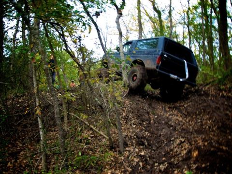 1995 Ford Bronco - A dark-colored SUV navigates a challenging, muddy off-road trail surrounded by dense trees.