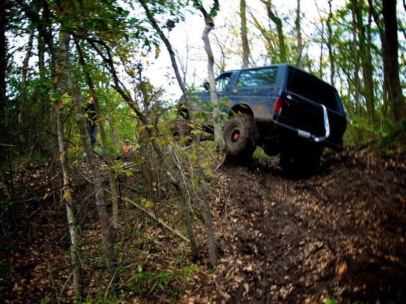 Black SUV driving up a muddy hill in a wooded area. Trees and foliage surround the trail