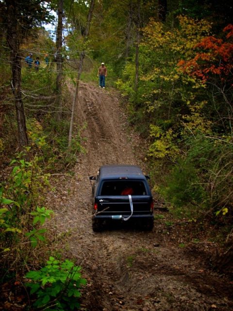 1995 Ford Bronco - A dark SUV navigates a steep, muddy off-road trail surrounded by autumn foliage.