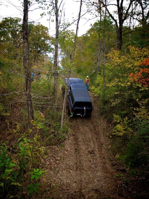1995 Ford Bronco - A dark 1995 Ford Bronco navigates a steep, muddy incline surrounded by trees in autumn.