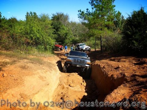 1995 Ford Bronco - Several off-road vehicles navigate a muddy trail under a clear blue sky.