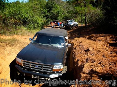 1995 Ford Bronco - A black 1995 Ford Bronco drives through a challenging dirt path, with several other off-road vehicles visible behind it.