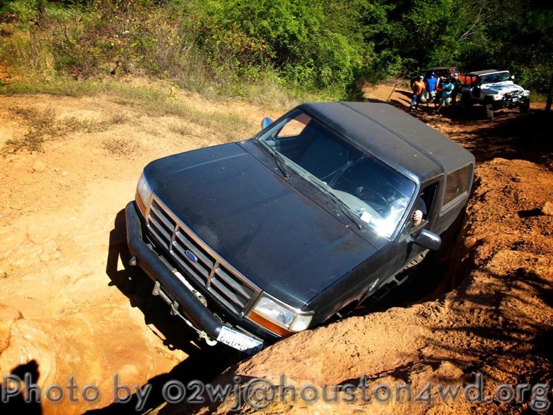 Dark 1995 Ford Bronco navigating a rutted dirt trail with other vehicles and people in the background