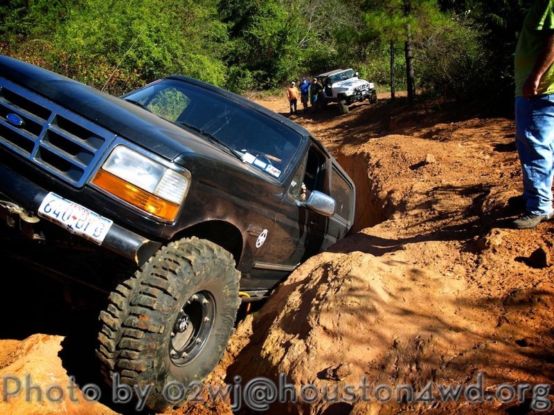 Black Ford Bronco navigating a deep rut on a dirt trail, with another white Jeep and people in the background