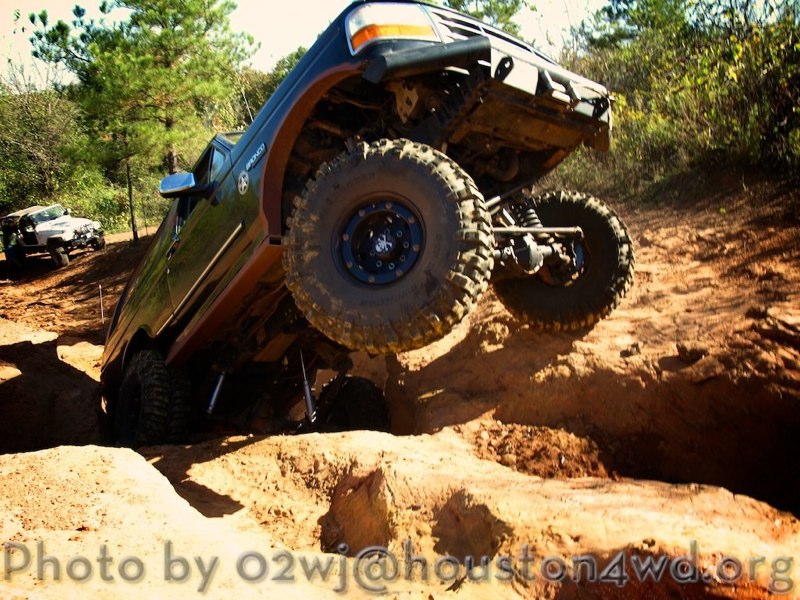 Dark blue Ford Bronco with lifted suspension and large tires climbing a dirt hill. Another white SUV is visible in the background