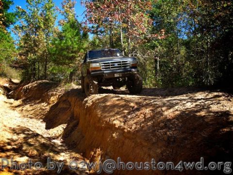 1995 Ford Bronco - A dark-colored Ford Bronco navigates a challenging off-road trail surrounded by trees.