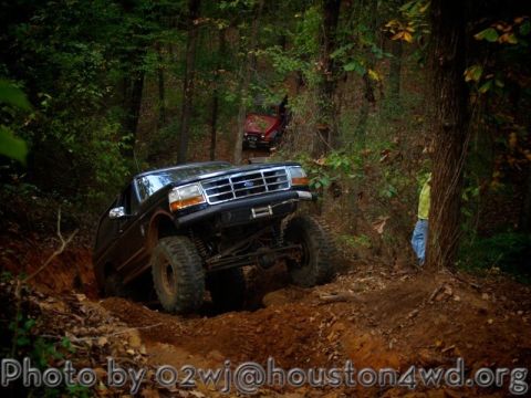 1995 Ford Bronco - A lifted black Ford Bronco navigates a challenging muddy trail in a wooded area, with another vehicle partially obscured in the background.