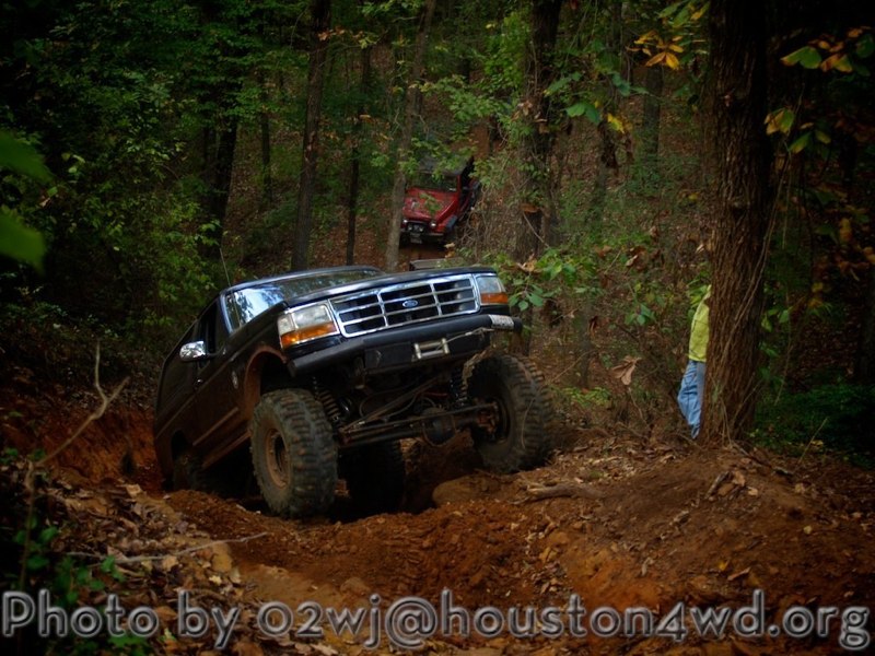 Black Ford Bronco on a muddy trail, lifted with large tires. Another red vehicle is visible behind it in the trees