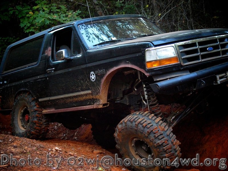 Black 1995 Ford Bronco with lifted suspension and large off-road tires driving on a muddy trail