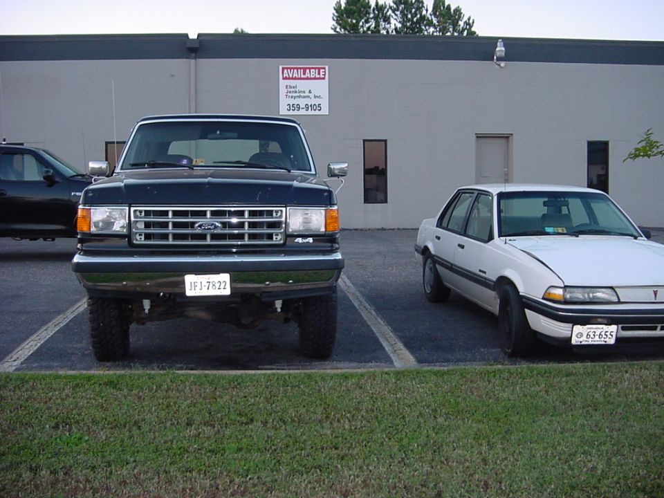 Dark green lifted Ford Bronco 4x4, front view, parked beside a white sedan at a commercial building