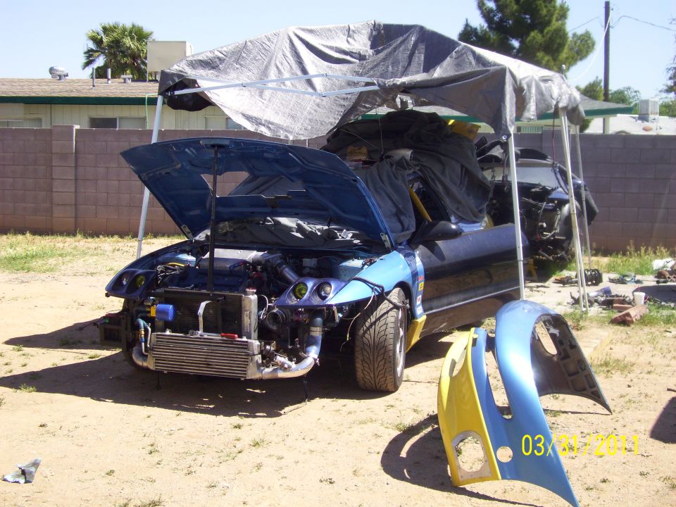 blue car with hood open, engine exposed, parts removed, parked under a tarp