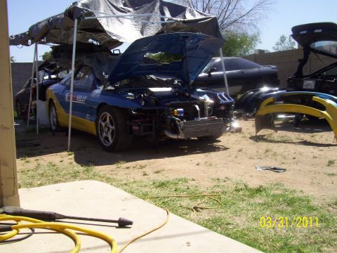 1995 Ford Probe - A blue and yellow car is partially disassembled under a tarp, with other car parts scattered around.