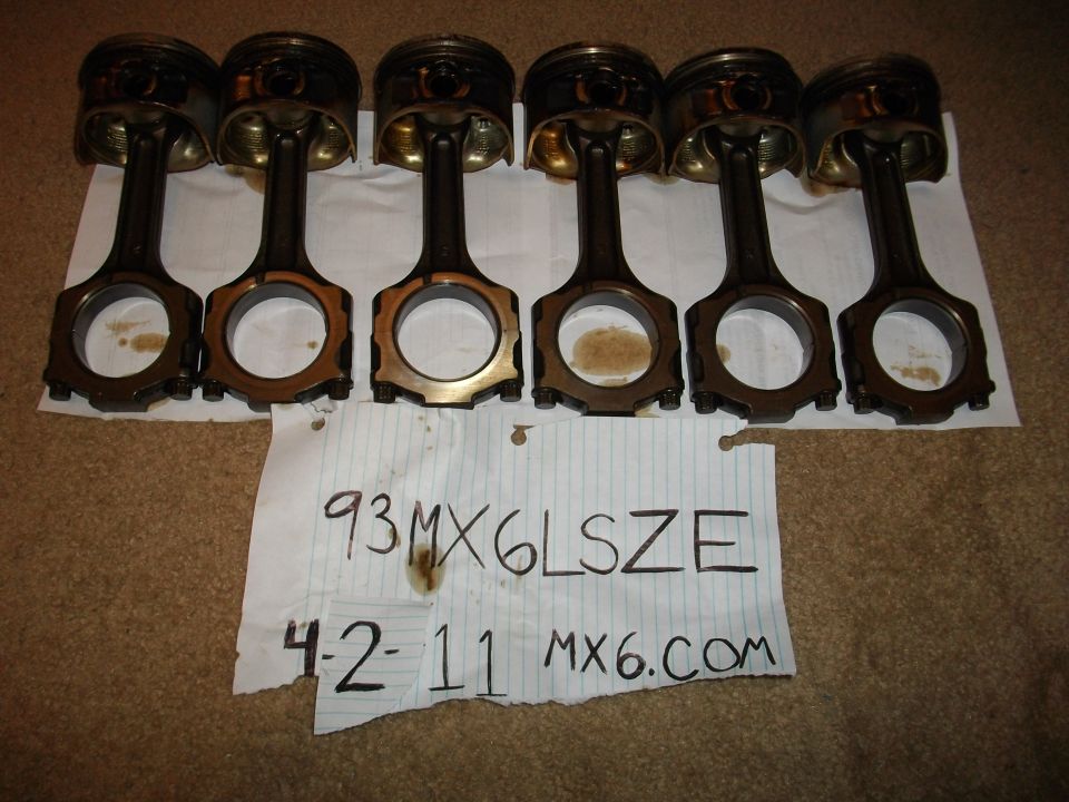 Six used engine connecting rods and pistons laid out on a carpeted surface