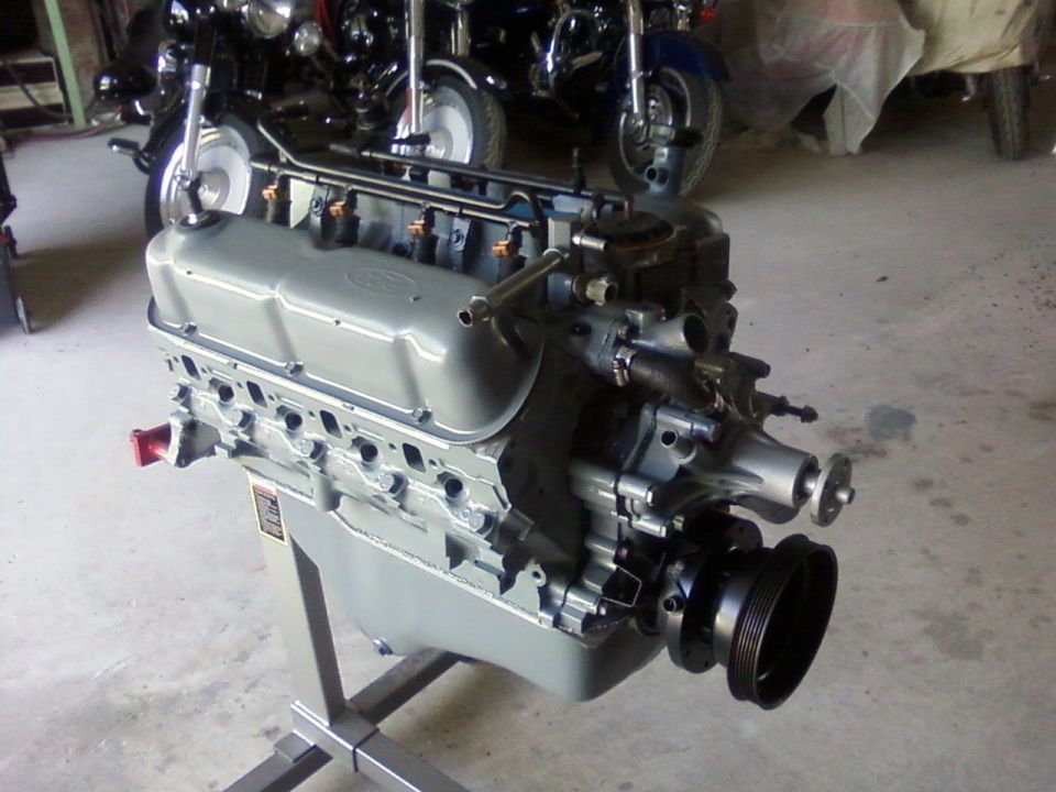 Gray Ford V8 engine on a metal stand, with motorcycles in the background