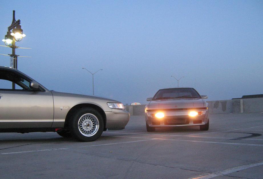 gold Mercury Grand Marquis is parked on the left, facing a silver sports car with its headlights on