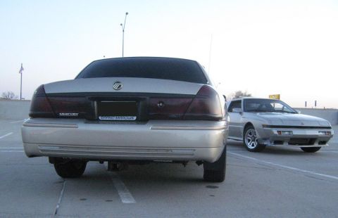 2000 Mercury Grand Marquis - A silver 2000 Mercury Grand Marquis is parked next to a silver sports car, viewed from the rear.