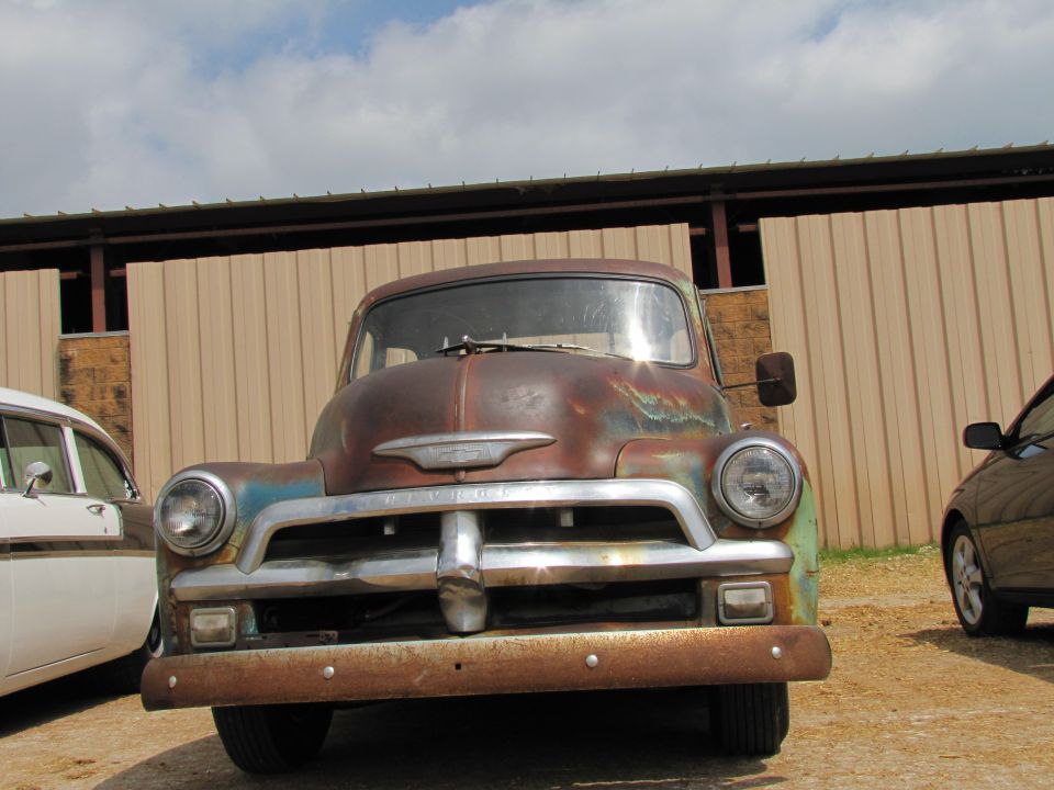 Front view of a rusty 1954 Chevrolet truck with a white car to the left and a dark car to the right