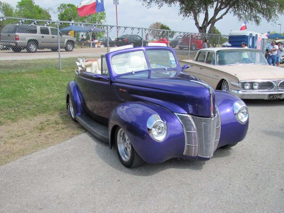 Purple convertible with white interior parked next to a tan car. Texas flag visible in background