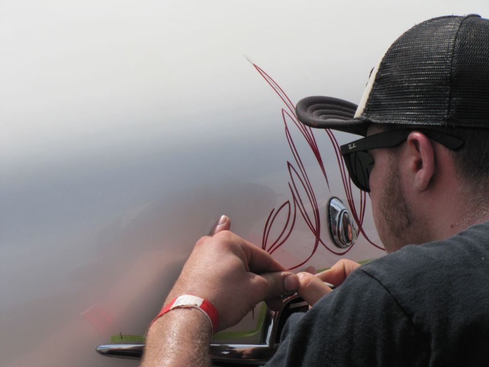Person wearing a trucker hat and sunglasses applies red pinstripes to a car's body panel
