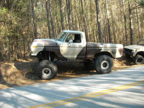 1979 Ford F-150 - A lifted pickup truck with a camouflage paint scheme sits parked on the shoulder of a road, with another vehicle visible behind it.