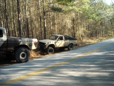 1979 Ford F-150 - Two trucks are parked on the shoulder of a road in a wooded area, with one truck partially obscuring the other.
