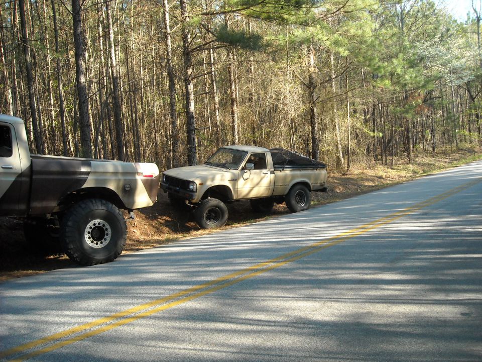 Tan Toyota pickup truck with black tonneau cover parked next to a camouflage Ford truck on a wooded roadside