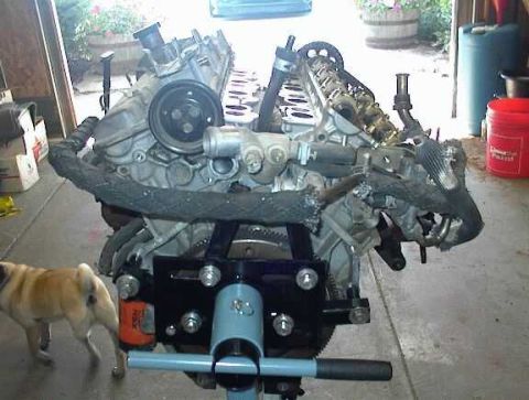 1997 Ford Taurus - A bare aluminum V8 short block mounted on a blue-and-black engine stand in a garage doorway, viewed from the transmission/bellhousing end w…