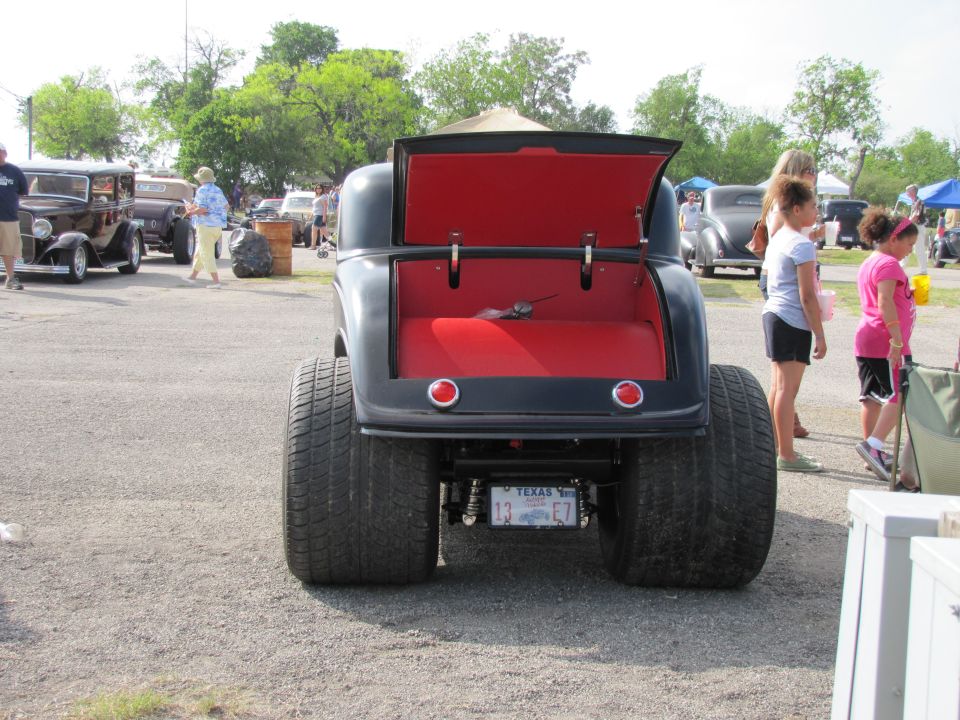 Rear view of a black hot rod with red interior and massive rear tires. Other cars and people are visible in the background