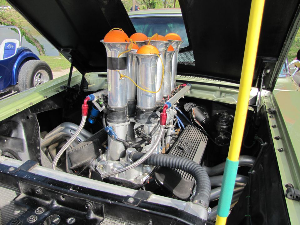 Custom V8 engine with chrome stacks and orange air filters, visible in a light green car. Another blue vehicle is partially visible