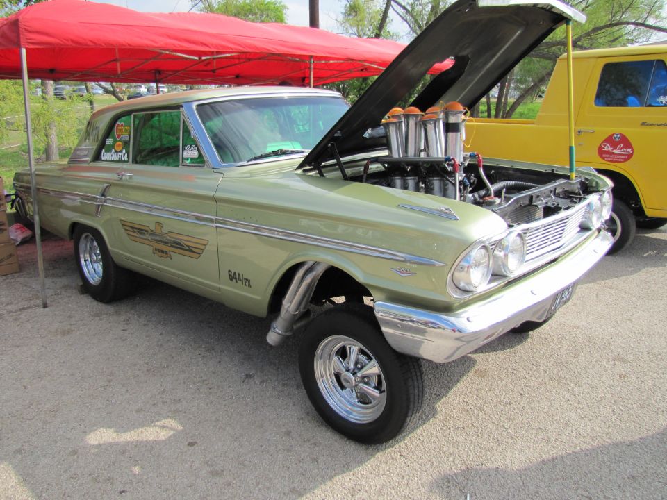 Light green vintage Ford Fairlane with hood open showing custom engine, drag racing setup, and chrome details. Yellow van parked behind