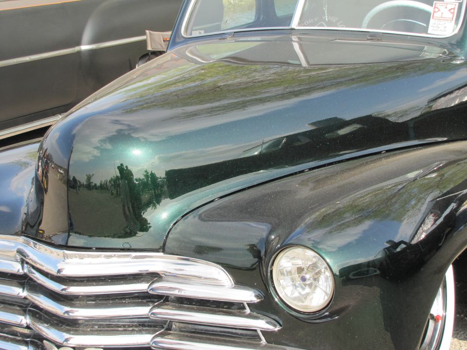 Green vintage car hood, chrome grille, and headlight. Another car is partially visible to the left