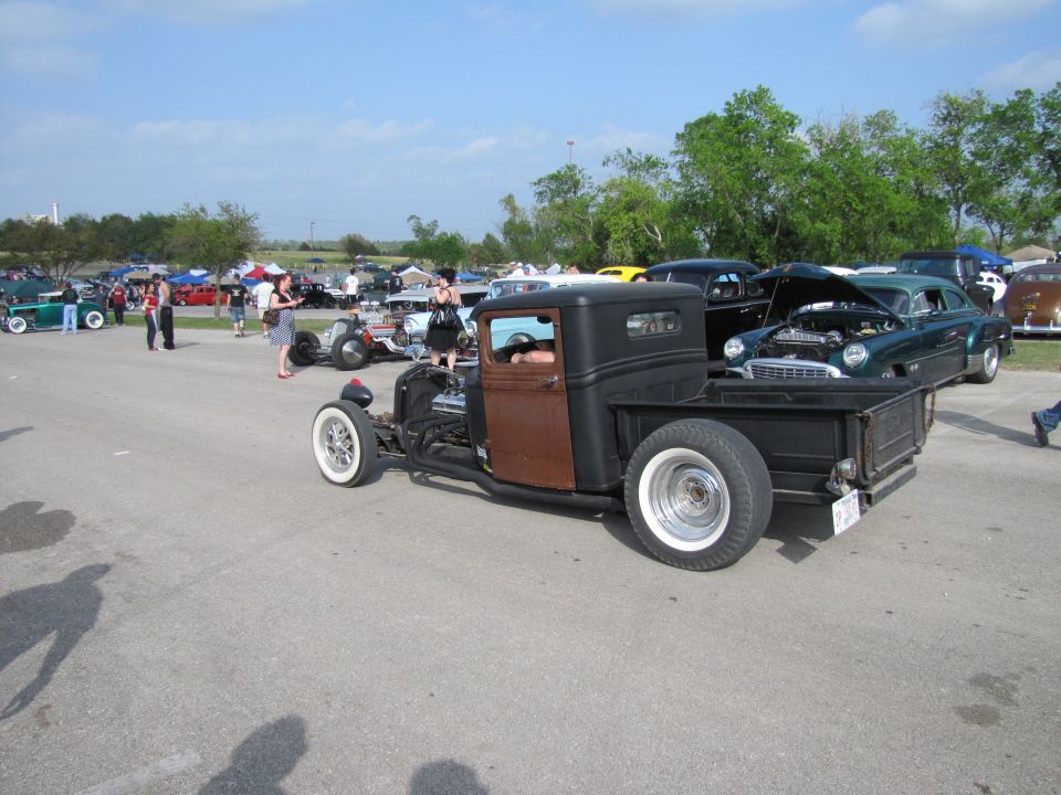 Hot rod pickup truck with exposed engine and wide whitewall tires, parked at an outdoor car show with other classic vehicles