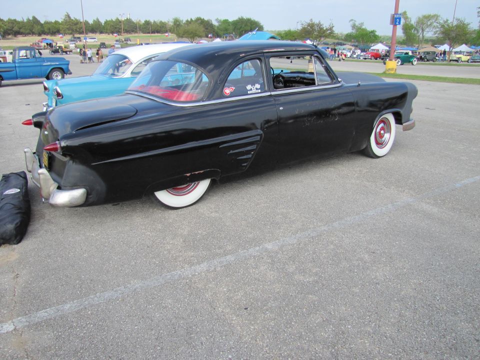 Black classic sedan with red interior, white wall tires, and chrome accents. Another car and a blue truck are visible in the background
