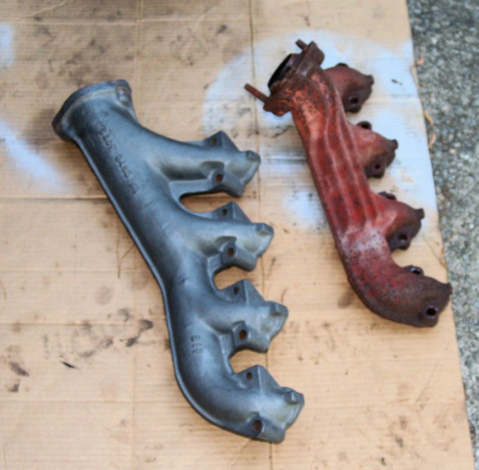 Two cast iron exhaust manifolds, one painted gray, the other rusted red, on a cardboard surface