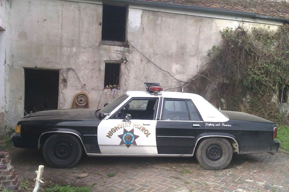 Black and white 1991 Ford Crown Victoria police car parked in front of a weathered building