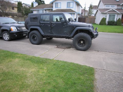 2008 Jeep Wrangler - Side profile of a lifted black four-door Jeep Wrangler JK Unlimited hardtop parked on a suburban driveway, riding on mud-terrain tires and…