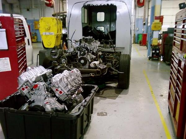 Vehicle chassis in shop with engines in a bin nearby. Another vehicle is to the left