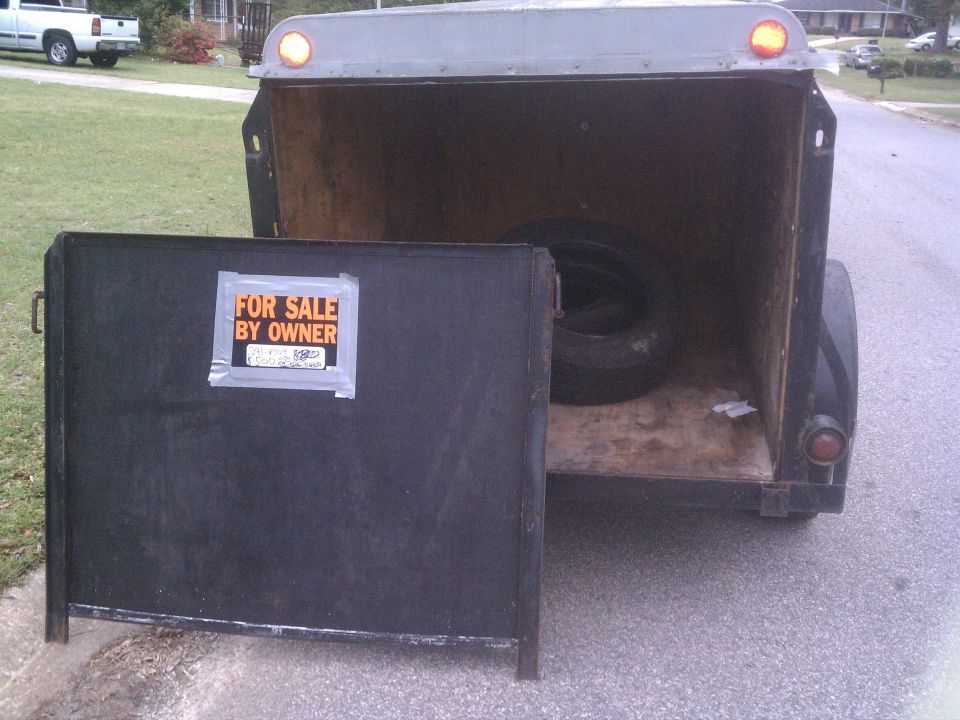 Trailer with open rear door and "FOR SALE BY OWNER" sign. Spare tire inside