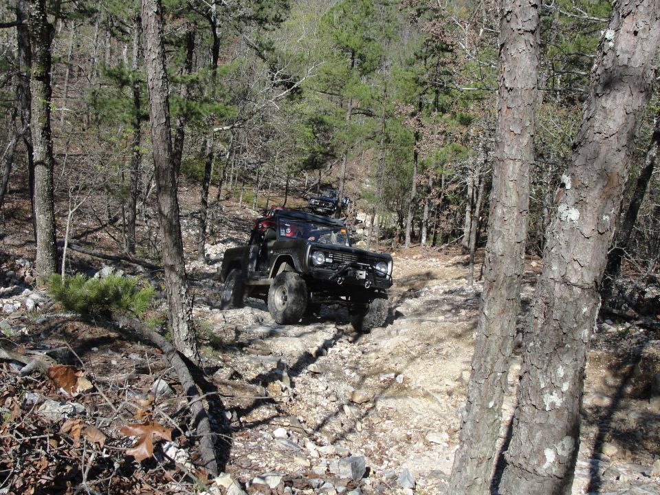 Off-road vehicle navigating a rocky trail through a wooded area. Another vehicle is visible behind it