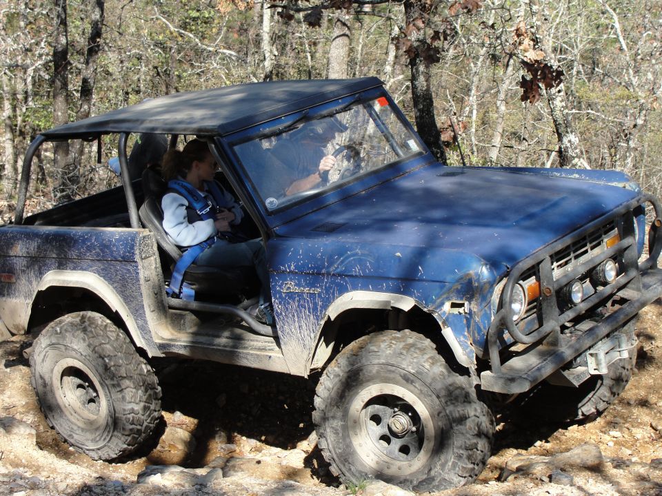 Blue early Ford Bronco with no doors and roll cage crawling rocks on a wooded trail