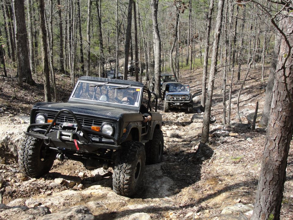 Black off-road vehicle with winch on rocky trail, another vehicle behind it in woods