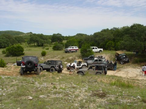 1973 Ford Bronco - A group of off-road vehicles gathered on a rocky, wooded terrain under a partly cloudy sky.