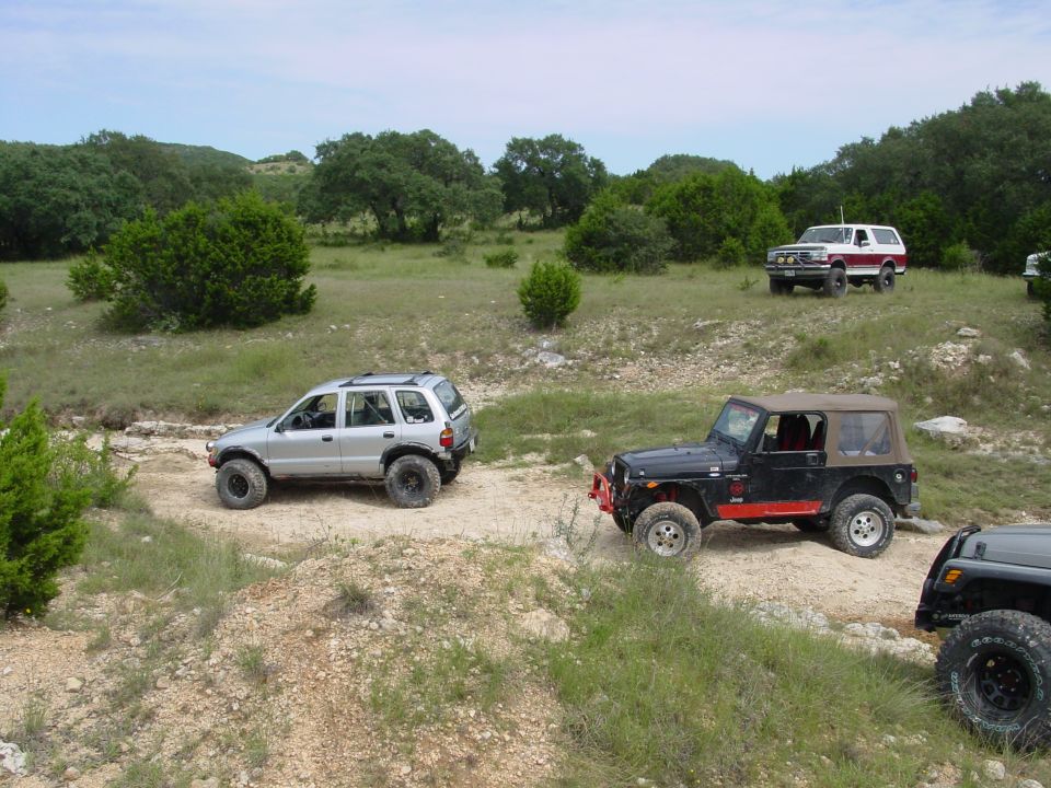 Several off-road vehicles, including a silver SUV and two Jeeps, on a dirt path in a grassy, wooded area