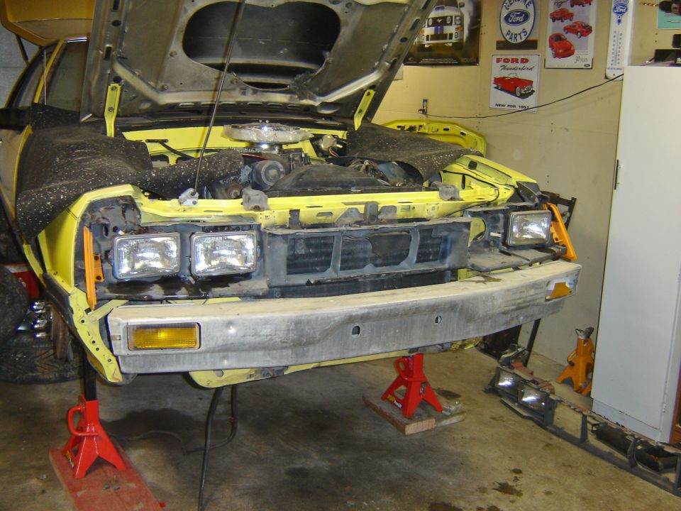 Yellow 1979 Ford Mustang front end on jack stands, hood open, bumper removed, headlights detached